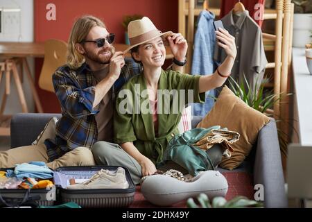 Glückliche junge Frau macht Selfie-Porträt auf ihrem Handy Zusammen mit ihrem Freund, während er mit Gepäck auf dem Sofa sitzt Stockfoto