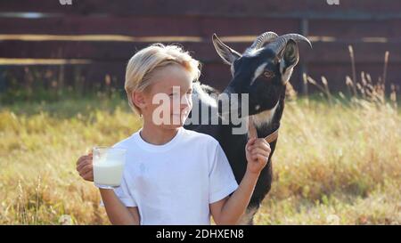 Ein Junge trinkt Ziegenmilch aus einem Becher neben seiner Ziege. Stockfoto