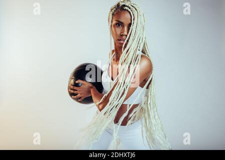 afroamerikanische Fitness-Frau mit langen geflochtenen Haaren hält Medizinball. Frau tut Training mit Medizinball auf weißem Hintergrund. Stockfoto
