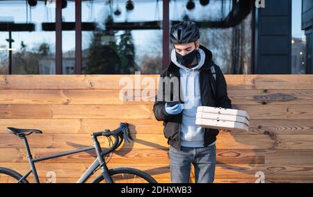 Fahrradlieferer im Helm hält ein Telefon und Pizzaboxen. Essen Lieferung, Essen zum Mitnehmen - Fahrrad Kurier Person bei der Arbeit im Freien Stockfoto