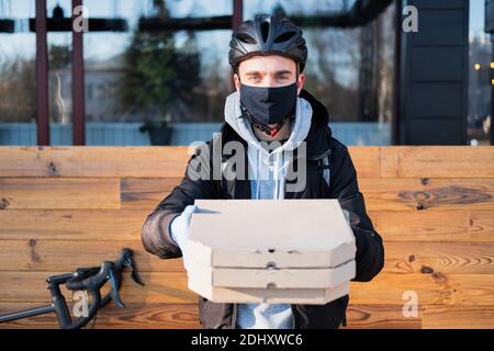 Der Lieferer hält Pizzataschachteln in einer ausgestreckten Hand, konzentriert sich auf das Gesicht. Lebensmittelzustellung, Fahrradkurier bei der Arbeit, Catering-Konzept zum Mitnehmen Stockfoto