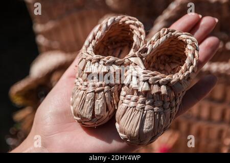 Souvenir Kinder Sandalen aus Stroh gewebt werden von einer weiblichen Hand gehalten. Kinderschuhe aus natürlichem Stroh handgefertigt auf der nationalen Handwerksmesse, i Stockfoto