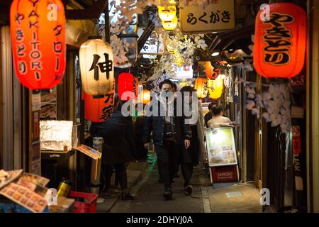 Besucher, die Gesichtsmasken tragen, gehen durch eine enge Straße eines Nachtbezirks in Shinjuku. Stockfoto