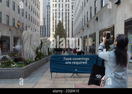 New York: Ein Tourist, der eine Maske trägt, fotografiert den Weihnachtsbaum des Rockefeller Center vor einem Schild mit der Aufschrift "Gesichtsbedeckung ist erforderlich". Stockfoto