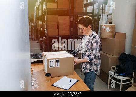 Beschäftigte Lagerarbeiterin, die Box mit Cello-Band verschließend, versiegelte Stockfoto