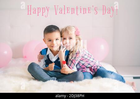 Alles Gute Zum Valentinstag. Weihnachtskarte mit Text. Zwei glückliche kaukasische Kinder essen herzförmige Lutscher. Junge und Mädchen feiern Valentinstag. Liebe Stockfoto