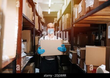 Verantwortlicher Lagerarbeiter, der Box trägt, Handschuhe und Maske trägt Stockfoto