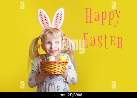 Frohe Ostern Karte mit Text. Kaukasische Mädchen in rosa Osterhasen Ohren halten Korb mit Eiern. Kind Kind im Studio auf gelbem Hintergrund. Komisches Kind c Stockfoto