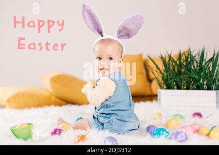 Frohe Ostern Karte mit Text. Kaukasische Baby Mädchen in lila Hase Ohren sitzen auf flauschigen weißen Teppich. Kind Kind spielt mit Ostern bunte Eier. Cel Stockfoto