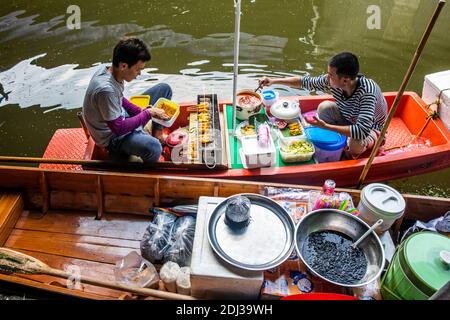 Zwei junge Männer in einer san-Pfanne, einer isst und ein anderer bereitet auf dem schwimmenden Markt in Damnoen Lebensmittel zu. Stockfoto