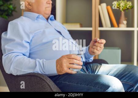 Senior Mann mit Glas Wasser Einnahme vorgeschriebene Pillen sitzen In einem Sessel zu Hause Stockfoto