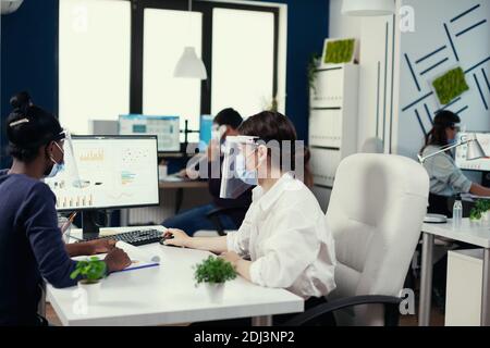 Diverse Kollegen, die auf Computer-Bildschirm mit wirtschaftlichen Graphen tragen Gesichtsmaske gegen covid19. Multiethnisches Team arbeitet in Unternehmen mit neuen normalen Respekt soziale Distanz während der globalen Pandemie mit Coronavirus. Stockfoto