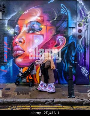 Zwei Frauen in Kopftüchern mit Kinderwagen laufen vor dem Street Art Wandbild von Mr. Cenz in der Bacon Street & Brick Lane, Shoreditch, East London, England Stockfoto