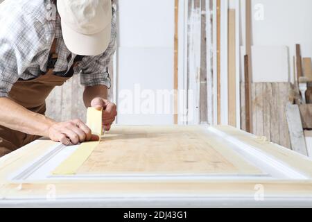 Männlicher Zimmermann, der das Holz in der Schreinerei bearbeitete, Papier-Abdeckband auf eine Holztür setzte, Overall und Kappe trug Stockfoto