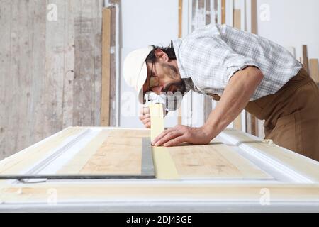 Männlicher Zimmermann, der das Holz in der Schreinerei bearbeitete, indem er Papier-Abdeckband auf die Holztür setzte, die der Linie des quadratischen Lineals aus Metall folgte und Ove trug Stockfoto