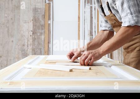 Männlicher Zimmermann, der das Holz in der Schreinerei arbeitet, Holzstäbchen auf eine Tür setzt und insgesamt trägt Stockfoto