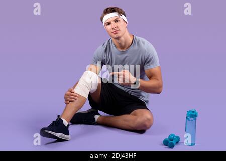 Athletic Millennial Mann mit Sportverletzung, zeigt auf seine verstauchten banded Knie über Flieder Studio Hintergrund Stockfoto