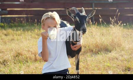 Der Junge trinkt Ziegenmilch aus einem Becher und umarmt seine geliebte Ziege. Stockfoto