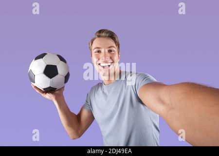 Lächelnder junger Mann mit Fußball nimmt Selfie auf seine Mobiltelefon über violettem Studiohintergrund Stockfoto