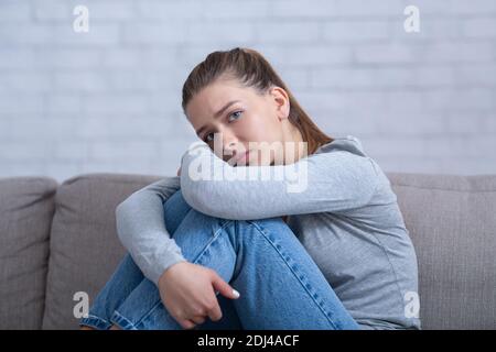 Junge depressive Frau umarmte ihre Knie auf dem Sofa zu Hause, in negative Gedanken absorbiert, mit psychischen Störungen Stockfoto