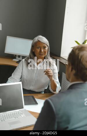 Zwei leitende Supervisoren unterhalten sich fröhlich miteinander und sitzen gegeneinander. Asiatische reife Geschäftsfrau im Gespräch mit einem Manager arbeiten auf Computer Stockfoto