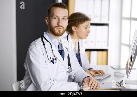 Rotbärtiger Arzt mit einer Kollegin, die die aktuelle Krankheitstherapie diskutiert, während sie am Arbeitsplatz in der Klinik sitzt. Teamarbeit in der Medizin Stockfoto