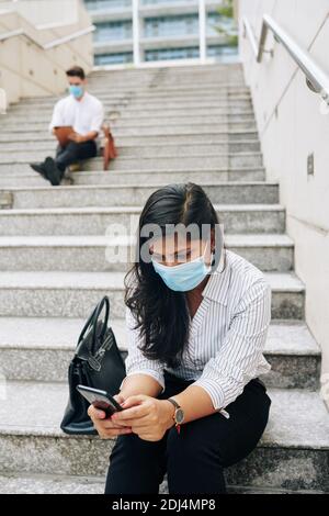 Serious junge indische Geschäftsfrau in medizinische Maske sitzt auf Stufen Und Textnachricht auf dem Smartphone-Bildschirm lesen Stockfoto
