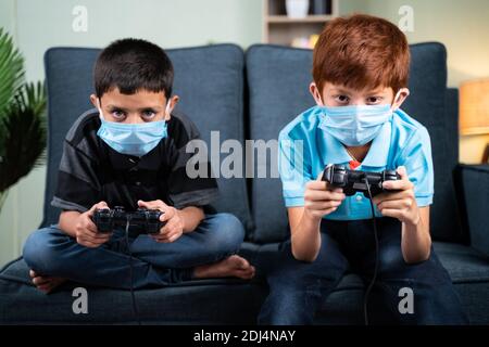 Zwei Kinder mit medizinischer Gesichtsmaske sind ernsthaft damit beschäftigt, Videos zu spielen Spiel mit Joystick zu Hause - Konzept der Kinder auf Spiel während Coronavirus covid-19 Lockdow Stockfoto