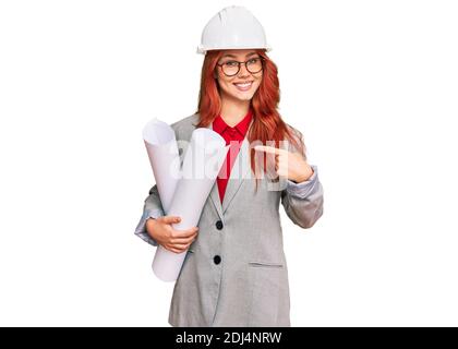 Junge rothaarige Architektin Frau trägt Schutzhelm mit Blaupausen lächelnd Glücklich zeigen mit Hand und Finger Stockfoto