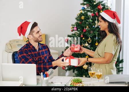 Lächelnde junge Mitarbeiter in den Händen des Weihnachtsmannes tauschen Weihnachtsgeschenke aus Im Büro Stockfoto