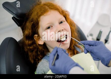 Ein Kind an der Zahnarztrezeption. Nahaufnahme Porträt von hübschen kleinen Mädchen mit roten lockigen Haaren, sitzen mit offenem Mund in Zahnarztstuhl, während Stockfoto