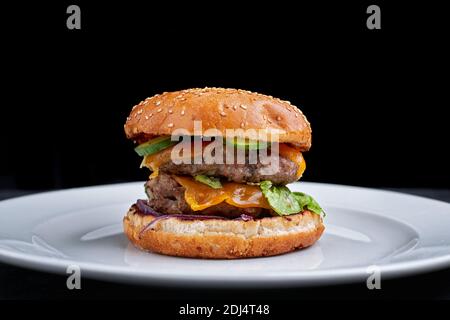 Burger mit doppeltem Fleischschnitzel, Käse. Gurke und Salat, auf weißem Teller, auf schwarzem Hintergrund Stockfoto