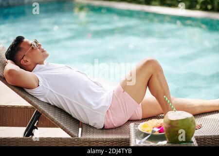 Fröhlicher junger Mann in Sonnenbrille, der beim Schwimmen auf der Chaiselongue ruht Pool mit Kokosnusscocktail und Obstteller auf dem Tisch In der Nähe Stockfoto