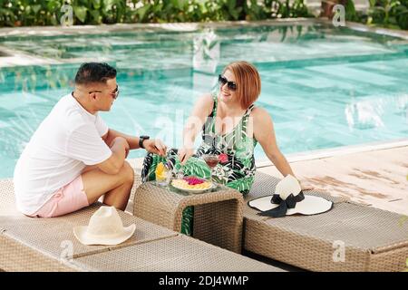 Fröhliches Paar in der Liebe, das am Pool ruht, Schnittfrüchte isst und Cocktails trinkt Stockfoto