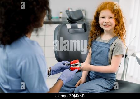 Glückliches lächelndes Kind im Zahnarztbüro. Kleines Mädchen mit roten lockigen Haaren, sitzt in Zahnarztstuhl und schaut in die Kamera, während ihre weibliche afrikanische Zahnärztin Stockfoto