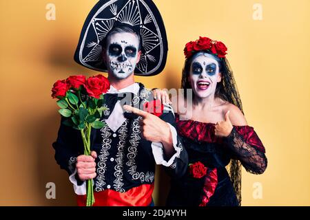 Junges Paar trägt mexikanische Tag der Toten Kostüm halten rosen lächeln glücklich zeigen mit Hand und Finger Stockfoto