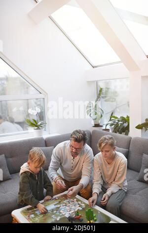 Vertikale Weitwinkel-Porträt von liebevollen Vater verbringen Zeit mit Zwei Söhne spielen Brettspiele und Puzzles zusammen in modern Inneneinrichtung Stockfoto