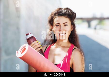 Porträt einer jungen Frau mit einem Turnteppich und einer Flasche Wasser in den Händen an einem sommerlichen sonnigen Morgen am Ufer. Stockfoto