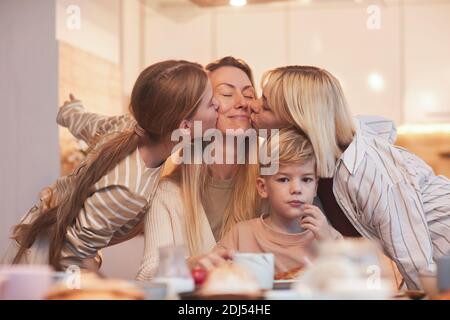 Portrait der glücklichen Mutter genießen Küsse von drei Kindern während Sitzen am Frühstückstisch in der Küche Stockfoto