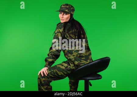 Foto von der Rückseite. Eine Frau in Militäruniform posiert auf einem schwarzen Stuhl. Grüner Hintergrund. Stockfoto