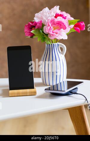 Ein Telefon, das mit einem kabellosen Ladegerät aufgeladen wird. Auf dem Tisch in der Wohnung neben der Blumenvase stehen Stockfoto