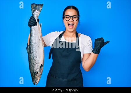 Junge schöne asiatische Mädchen Fischhändler Verkauf frische rohe Lachs zeigt Daumen nach oben zur Seite lächelnd mit offenem Mund Stockfoto