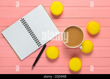 Ein Notizbuch mit einem Stift, einer Tasse Kaffee und gelben Makronen auf einem rosafarbenen Holztisch. Draufsicht. Flach liegend. Stockfoto