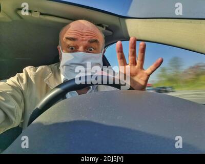 Senior mit Gesichtsmaske und erhobener Hand am Steuer eines PKW. Stockfoto