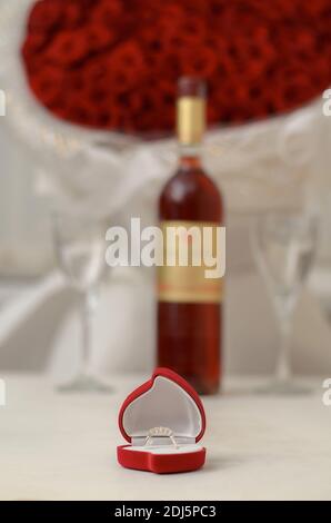 Silberner Ring in einer roten Schachtel in Herzform auf einem weißen Tisch auf dem Hintergrund einer Flasche Wein, leere Gläser und ein großes Bouquet von roter Rose Stockfoto