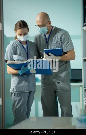Zwei moderne Kliniker in Uniform, Schutzmasken und Handschuhen diskutieren medizinische Dokumente, während einer von ihnen Notizen macht Stockfoto