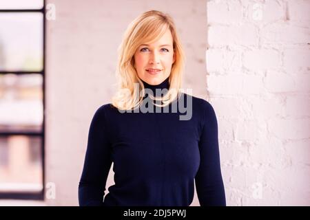 Porträtaufnahme einer schönen blonden Frau, die in die Kamera schaut und lächelt, während sie drinnen steht. Stockfoto