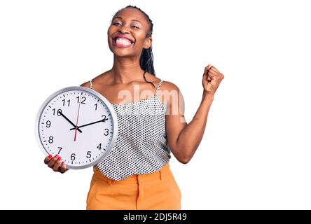 Junge afroamerikanische Frau, die große Uhr hält und stolz schreit, den Sieg und den Erfolg mit erhobenen Armen sehr aufgeregt feiert Stockfoto