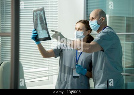 Glatze männlichen Kliniker in Uniform und Schutzmaske und Handschuhe Zeigt auf die Röntgenaufnahme der Lunge, die von seinem Assistenten während der Diskussion gehalten wird Von Pathologien Stockfoto