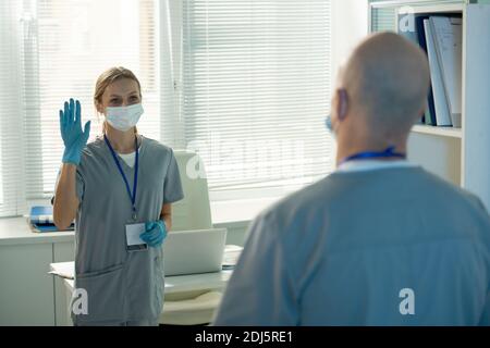 Junge Arbeiterin des medizinischen Labors in schützender Arbeitskleidung winken Hand zu kahlen männlichen Kollegen, während Begrüßung ihn in der Morgen Stockfoto